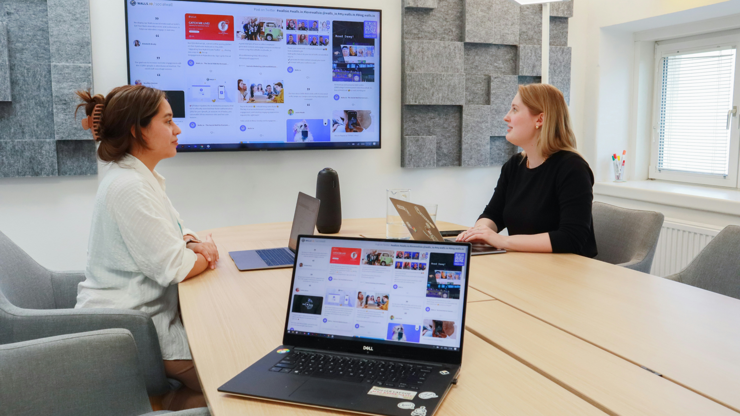 Two colleagues discussing a social media wall dashboard displayed on laptop and screen in a conference room