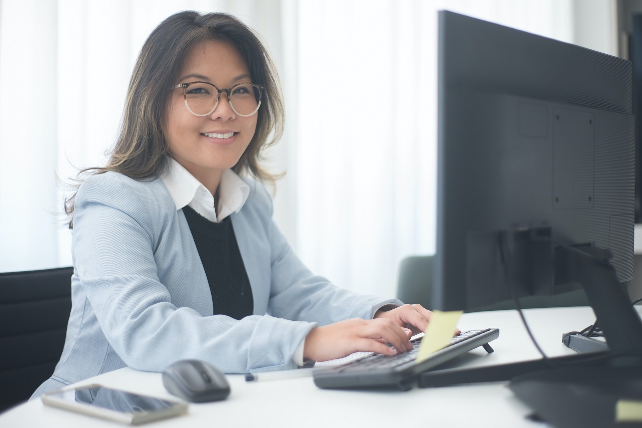 Smiling woman in glasses and a light blue blazer typing at a desktop computer in a bright office