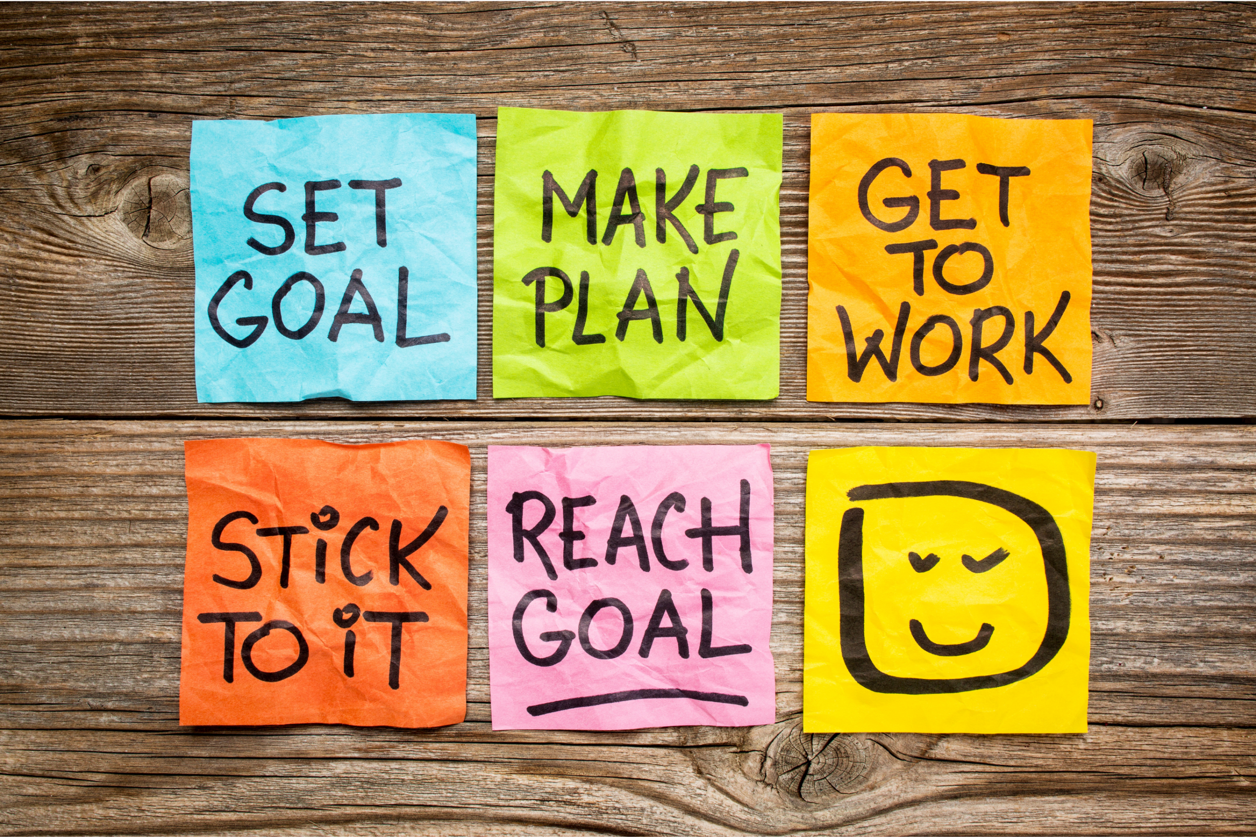 Six colourful sticky notes on wood reading "Set goal," "Make plan," "Get to work," "Stick to it," "Reach goal," and a smiley face