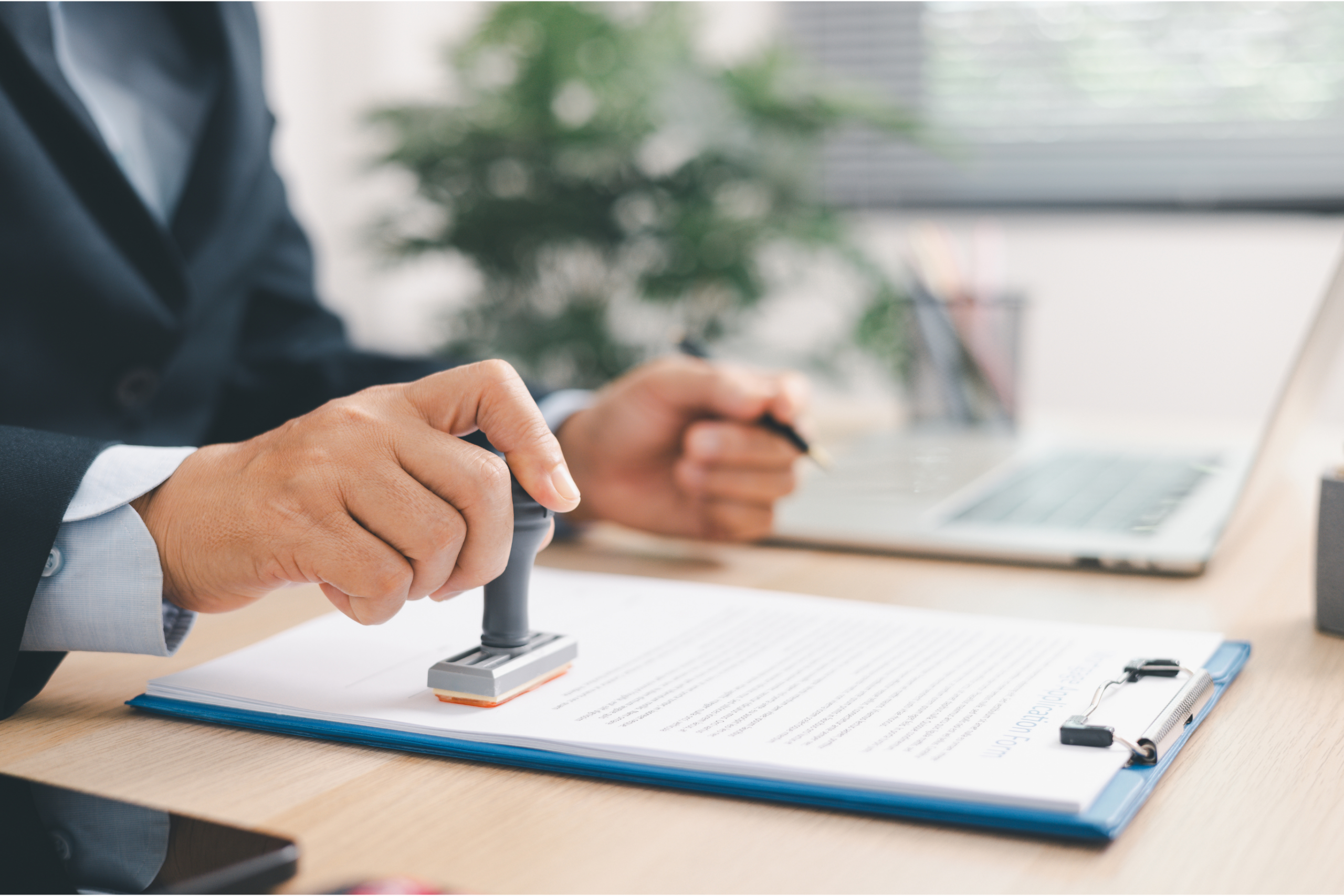 Person in a suit pressing a rubber stamp onto an application form on a blue clipboard, with a laptop on the desk behind