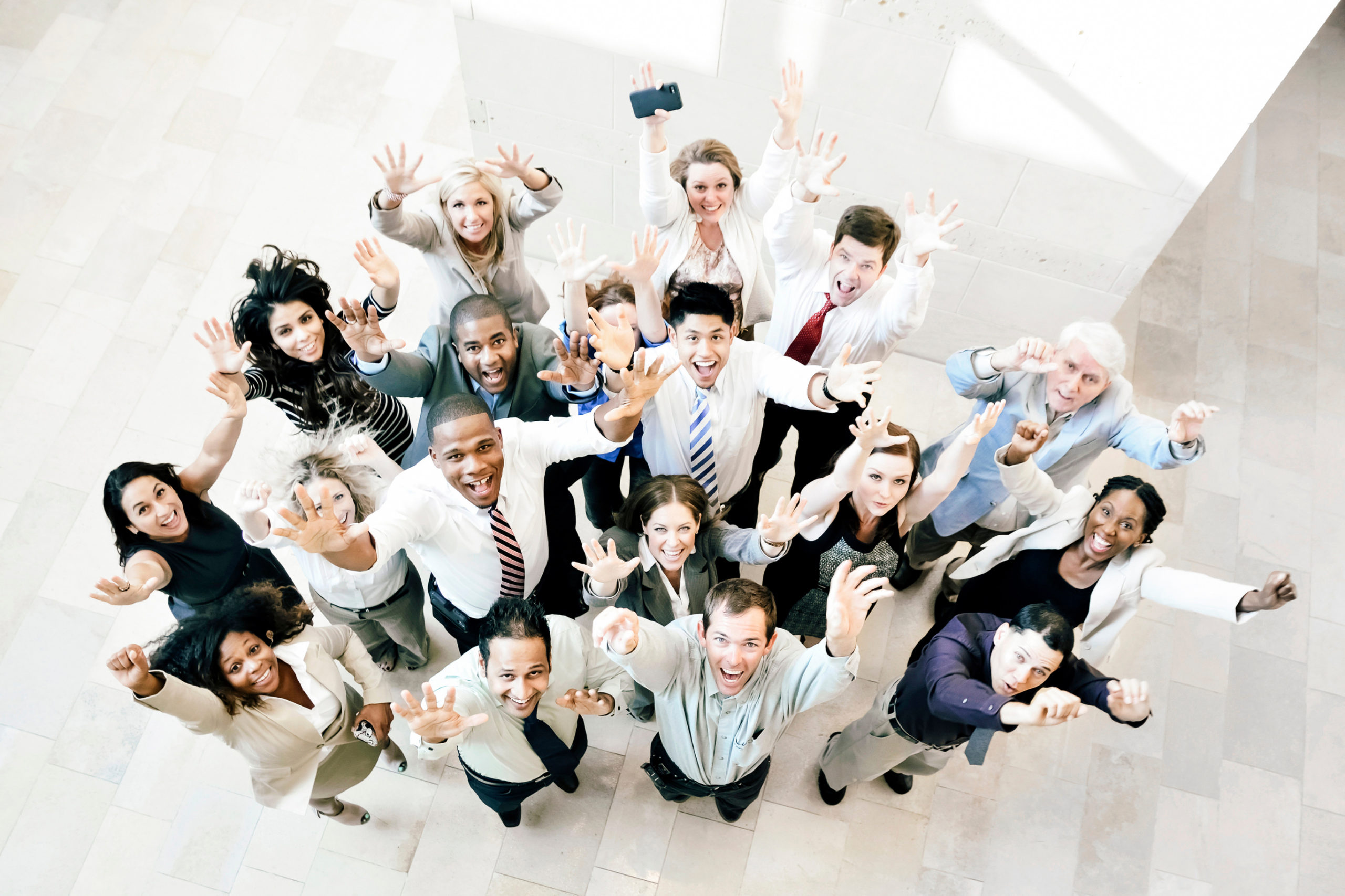 Large group of smiling professionals seen from above, waving and raising their arms toward the camera on a light tiled floor