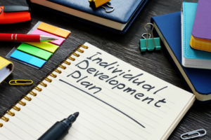 Individual Development Plan Notebook – Sincron HR Open spiral notebook with "Individual Development Plan" handwritten on the page, beside a marker, sticky notes, and stacked notebooks