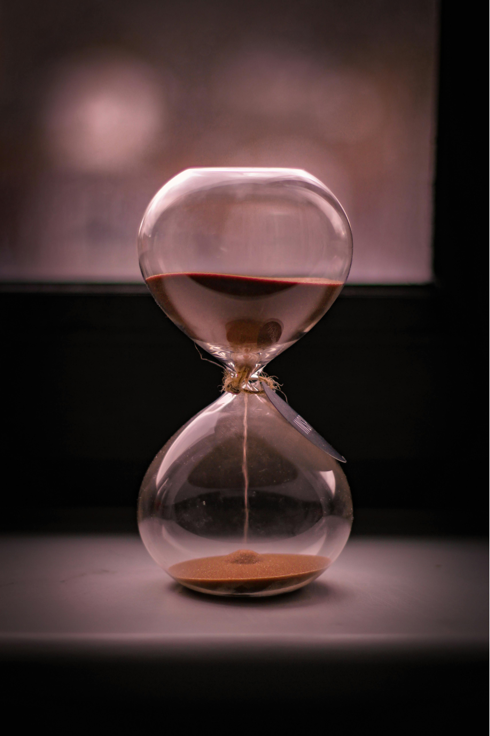 Hourglass with Falling Sand – Sincron HR Glass hourglass with sand running from the top bulb to the bottom, on a windowsill in soft light
