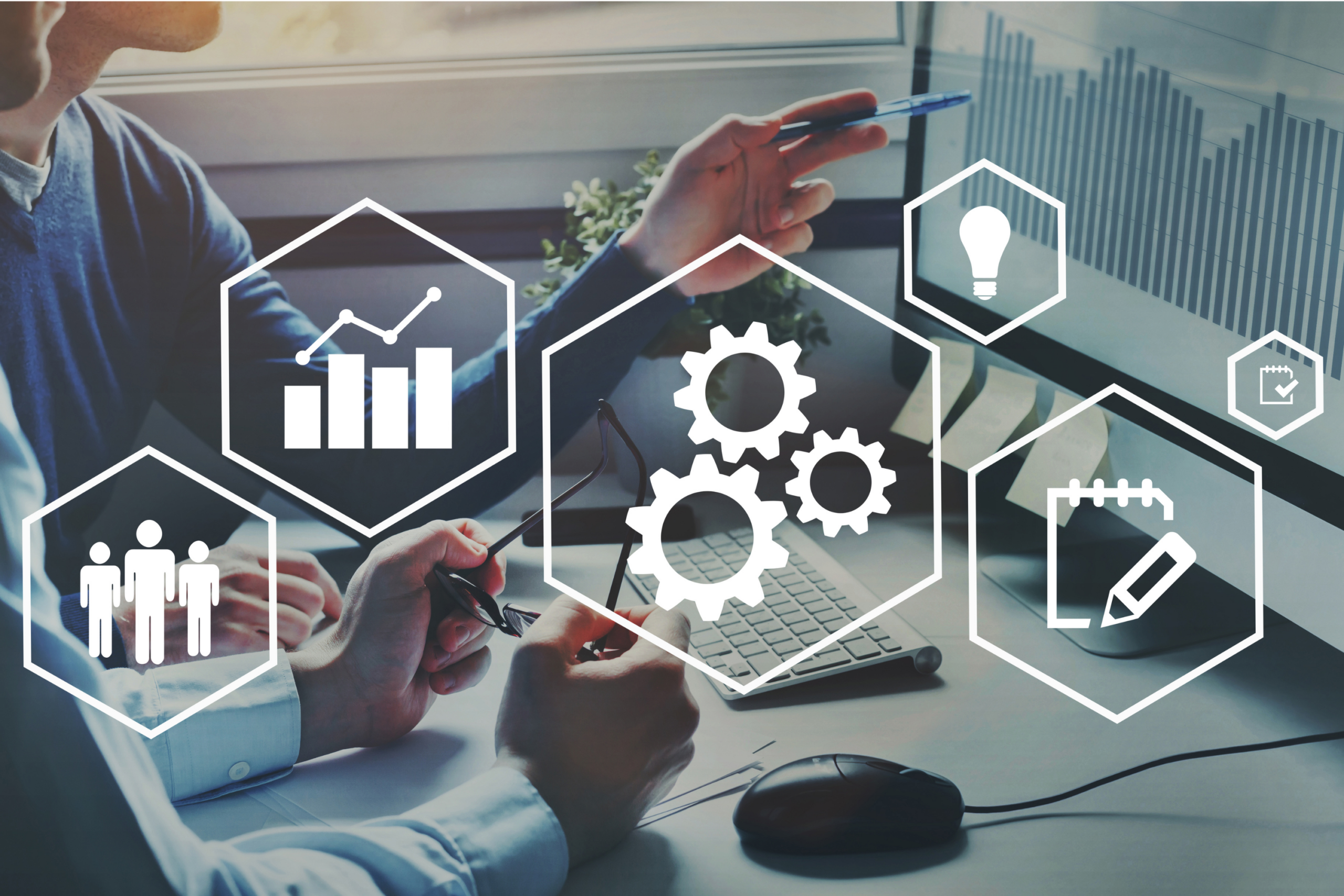HR Data and Planning Icons Over Office Desk – Sincron HR Two colleagues reviewing data at a desk, overlaid with hexagon icons of people, a bar chart, gears, a lightbulb, a notepad, and a checklist
