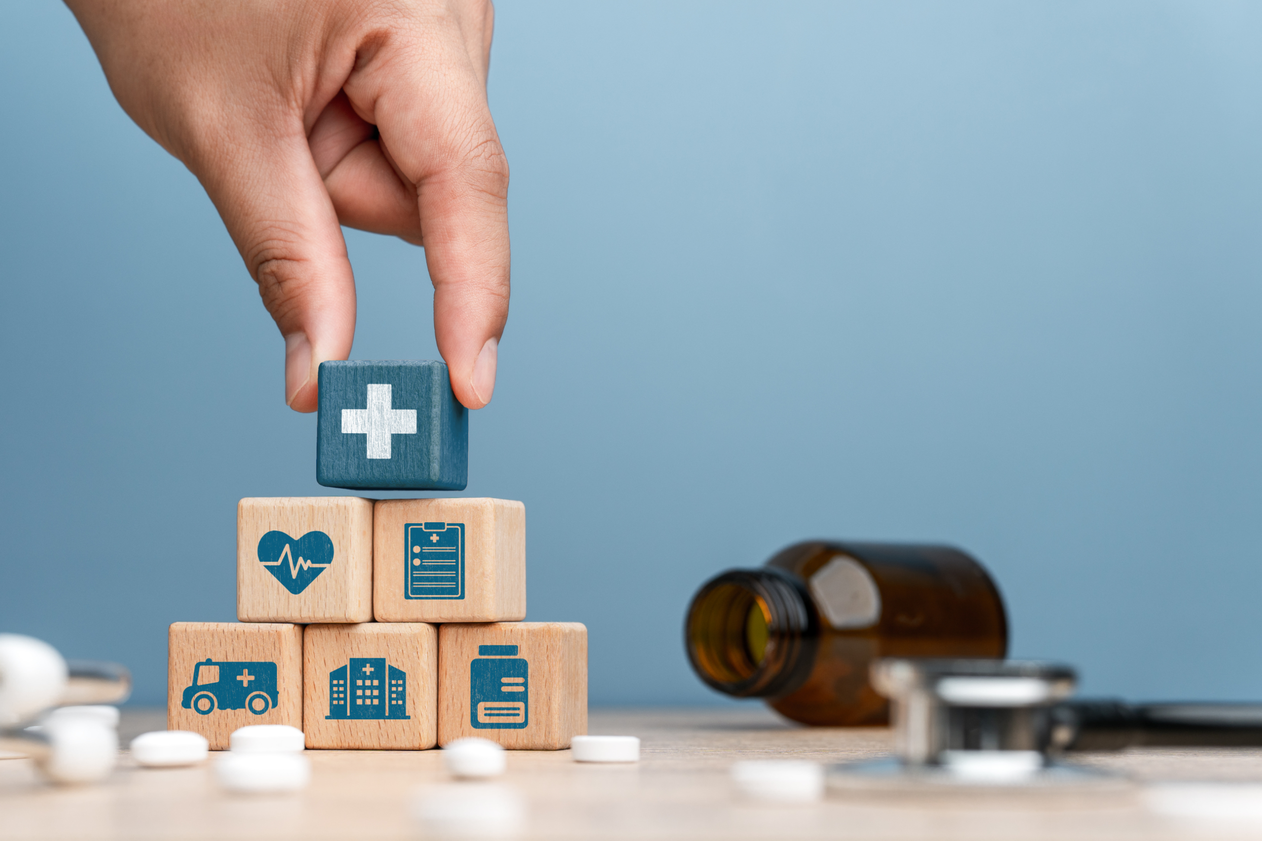 Healthcare Icon Blocks – Sincron HR Employee Benefits Hand placing a blue block with a white cross on top of wooden blocks showing a heart rate, medical clipboard, ambulance, hospital, and medicine bottle, with pills and a stethoscope nearby