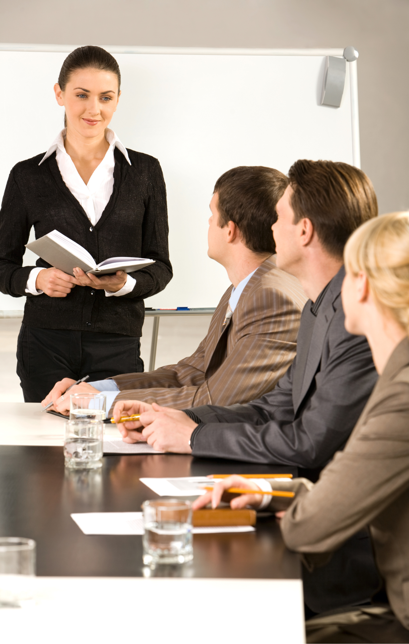 Presenter Leading a Meeting – Sincron HR Training Woman holding an open notebook presenting to three colleagues seated at a conference table, with a whiteboard behind her