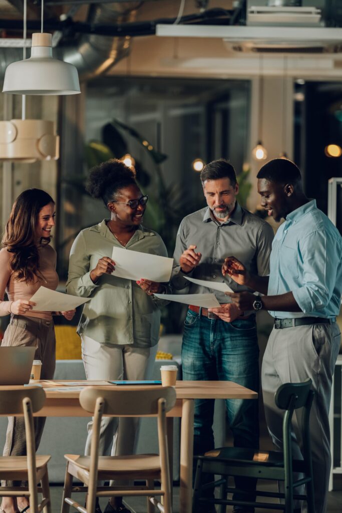 Four smiling colleagues standing by a table in a warmly lit office, sharing and discussing printed papers