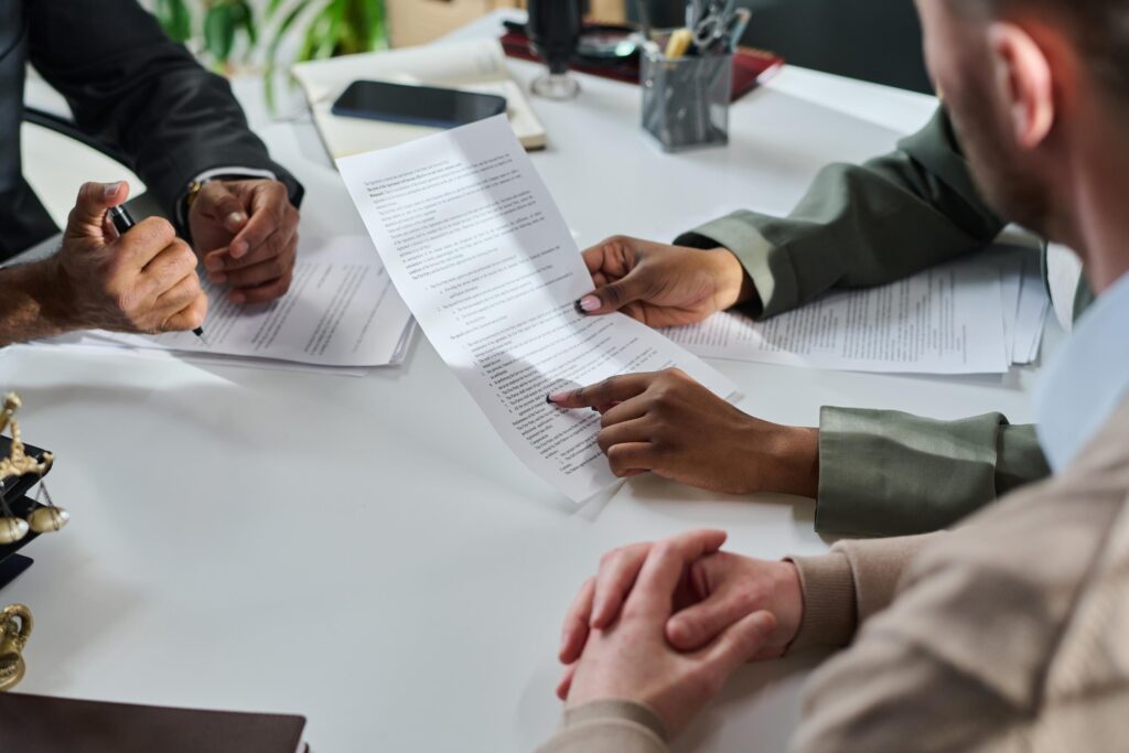 Close-up of three people at a white desk, one pointing to a clause in a printed agreement while another holds a pen, with small brass scales of justice nearby