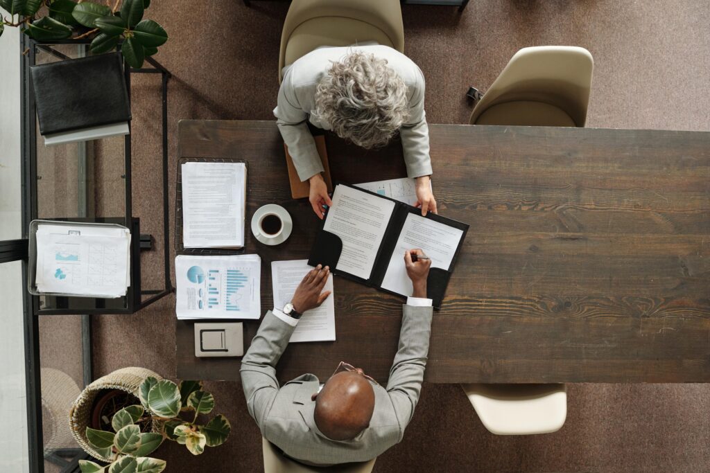 Overhead view of two people at a wooden table, one signing a document in a black folder held open by the other, with printed charts and coffee nearby